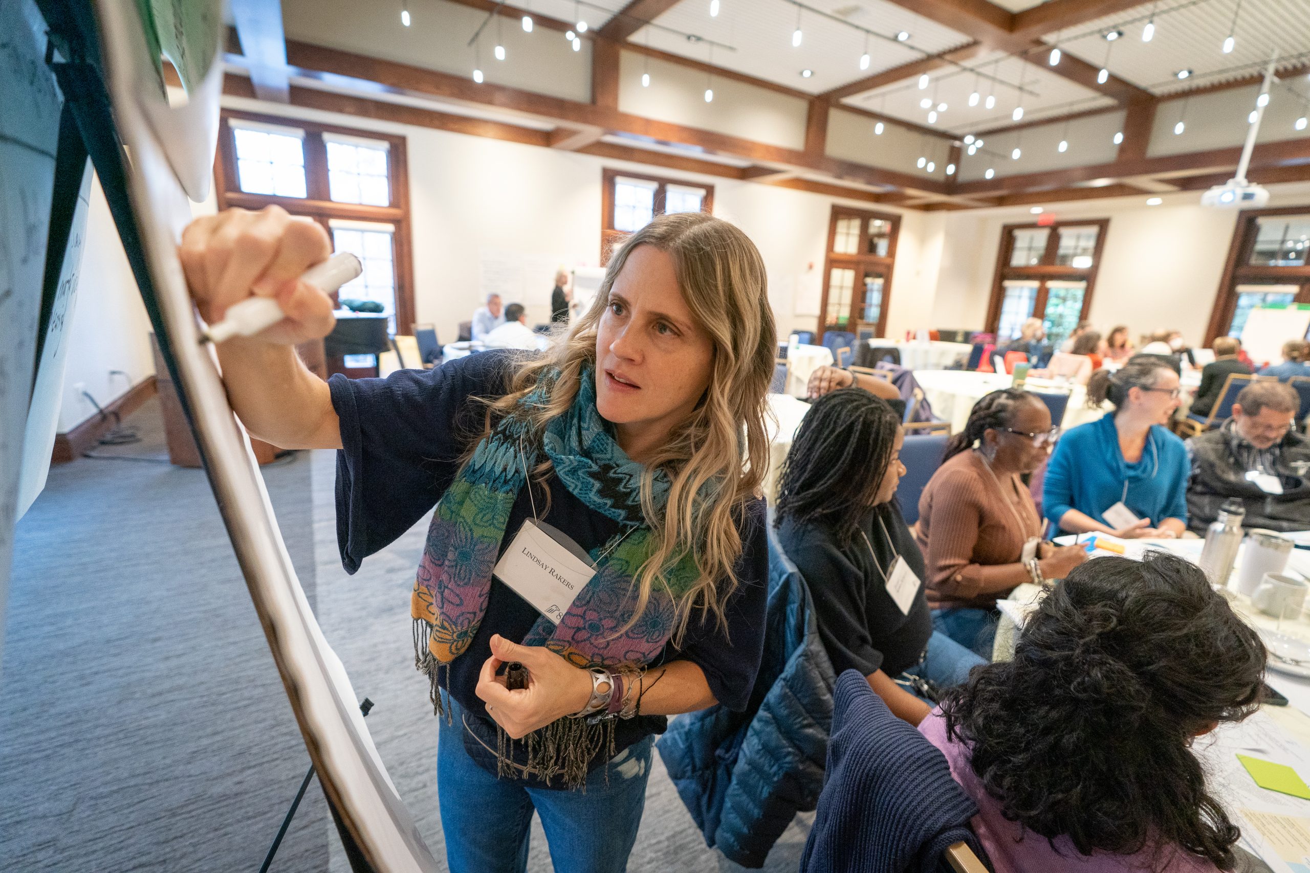 A woman draws on a white board in a full conference room.