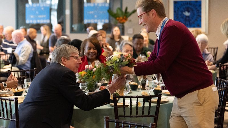 A man hands another some flowers at a luncheon.