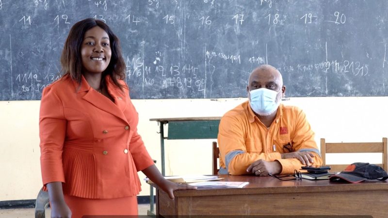 A woman in and orange dress stands by a desk.